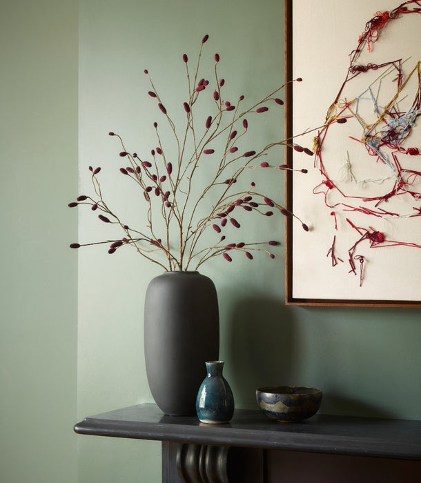 A dark grey matte ceramic vase filled with artificial pimpernel spray with deep burgundy buds, styled on a dark mantelpiece against a sage green wall next to a framed abstract painting.