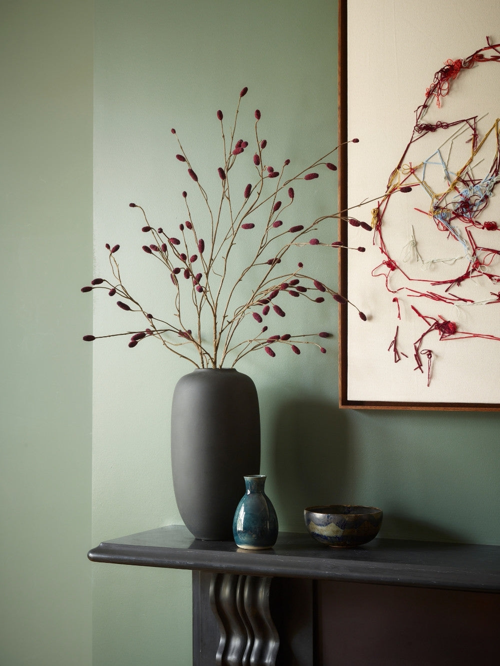 A dark grey matte ceramic vase filled with artificial pimpernel spray with deep burgundy buds, styled on a dark mantelpiece against a sage green wall next to a framed abstract painting.