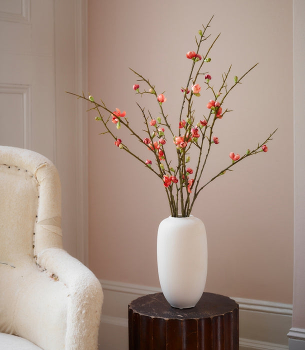A white ceramic vase filled with artificial quince blossom spray, featuring small orange / pink flowers, styled on a dark wood pedestal next to a cream-colored armchair against a beige wall.