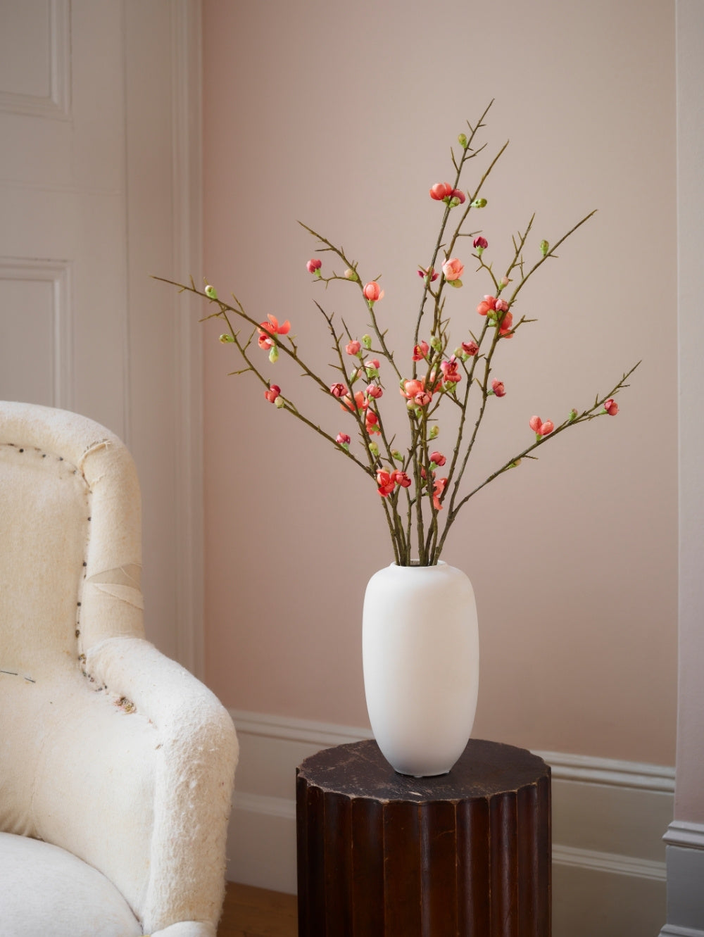 A white ceramic vase filled with artificial quince blossom spray, featuring small orange / pink flowers, styled on a dark wood pedestal next to a cream-colored armchair against a beige wall.