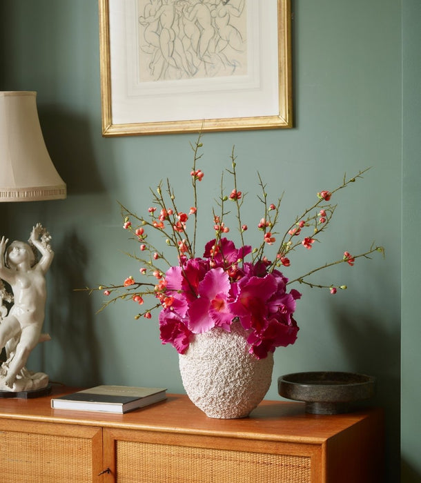 A textured white ceramic vase filled with bright magenta Vanda Orchid flowers and a delicate quince blossom spray with orange / pink flowers, styled on a wooden cabinet against a sage green wall with framed art.