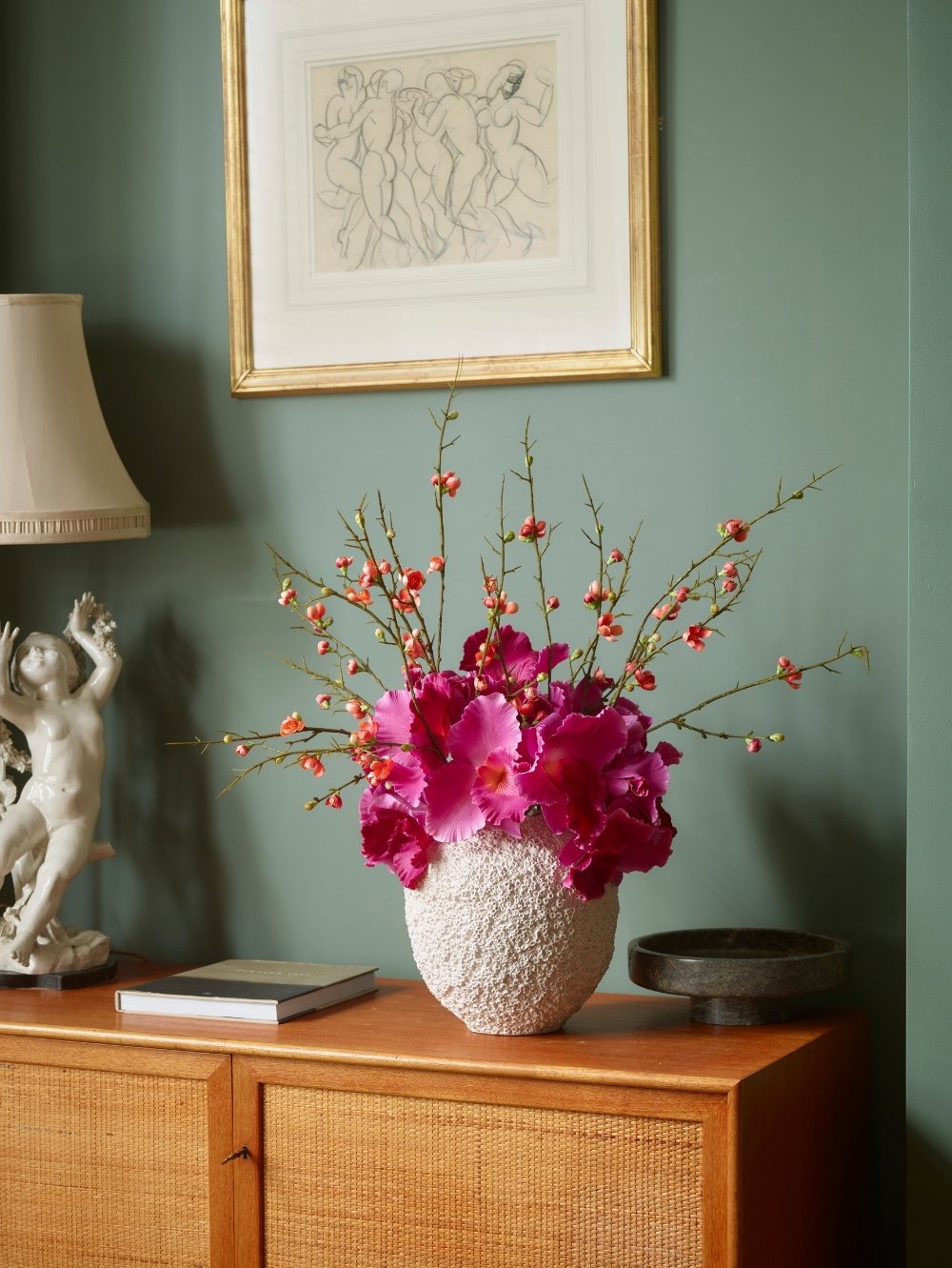 A textured white ceramic vase filled with bright magenta Vanda Orchid flowers and a delicate quince blossom spray with orange / pink flowers, styled on a wooden cabinet against a sage green wall with framed art.