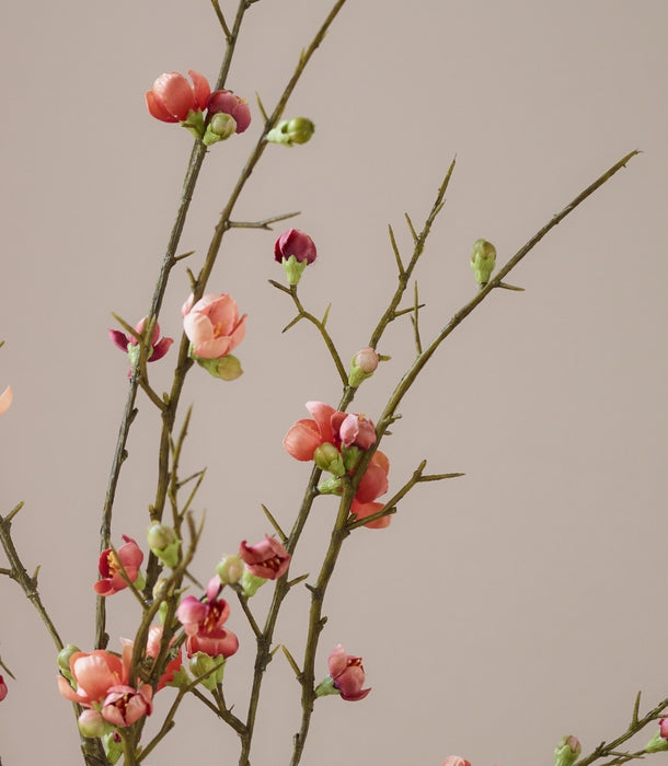 Close-up of hyper-realistic artificial quince blossom spray, featuring small orangey-pink flowers, green buds, and thorny brown stems against a solid warm beige background.