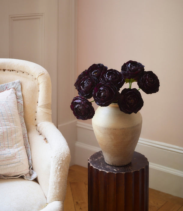 A ceramic vase filled with artificial dark burgundy ranunculus stems, displayed on a dark wood pedestal in a softly lit room next to a cream-colored armchair.