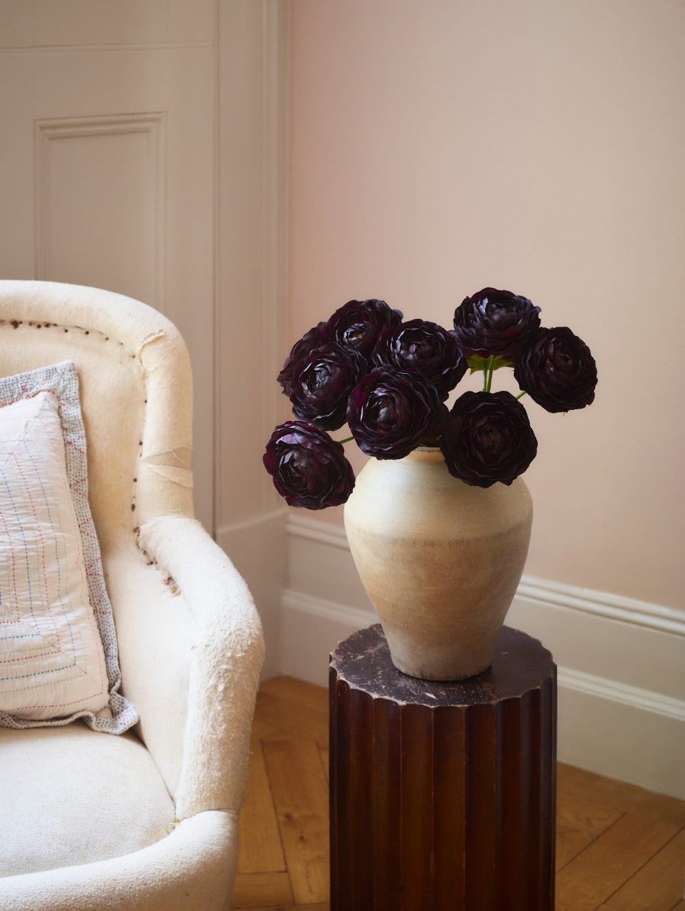 A ceramic vase filled with artificial dark burgundy ranunculus stems, displayed on a dark wood pedestal in a softly lit room next to a cream-colored armchair.