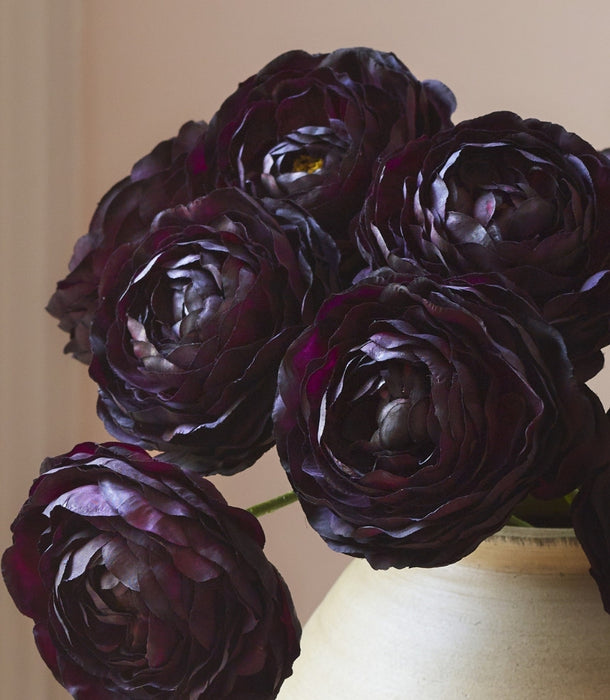 Close-up of hyper-realistic artificial dark burgundy ranunculus stems, arranged in a textured cream ceramic vase against a soft beige background.
