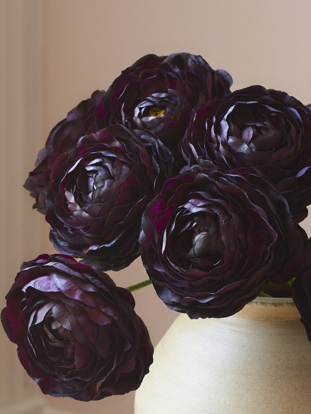 Close-up of hyper-realistic artificial dark burgundy ranunculus stems, arranged in a textured cream ceramic vase against a soft beige background.
