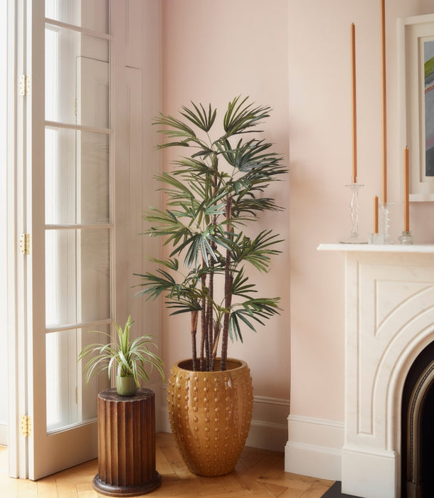 A tall potted artificial Raphis Palm in a textured gold pot stands beside a smaller potted spider plant on a ribbed wooden pedestal, set in a softly lit room with pale pink walls, wood floors, and a white fireplace.
