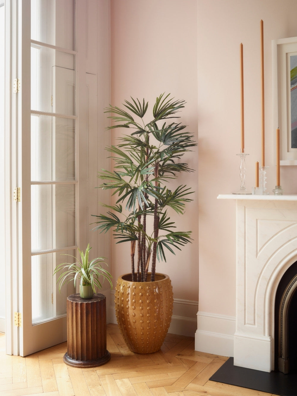 A tall potted artificial Raphis Palm in a textured gold pot stands beside a smaller potted spider plant on a ribbed wooden pedestal, set in a softly lit room with pale pink walls, wood floors, and a white fireplace.