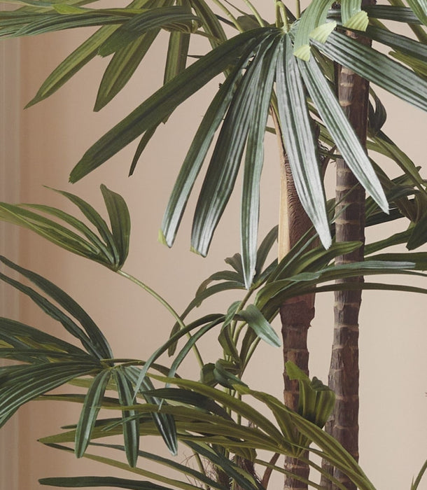 Close-up of a hyper-realistic artificial Raphis Palm with long green fan leaves and brown textured trunks against a plain beige wall.