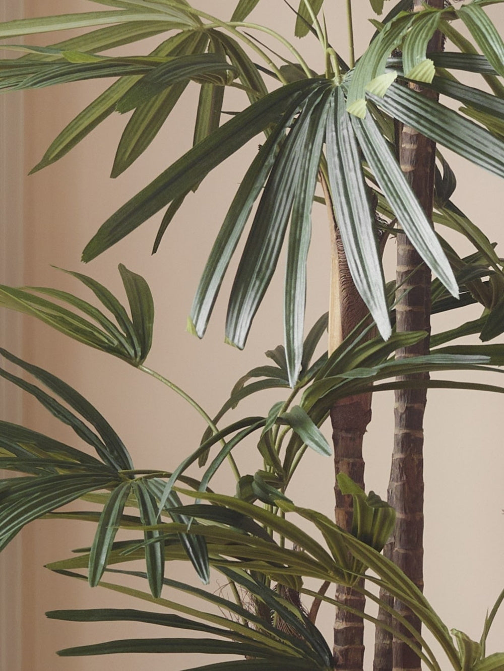 Close-up of a hyper-realistic artificial Raphis Palm with long green fan leaves and brown textured trunks against a plain beige wall.