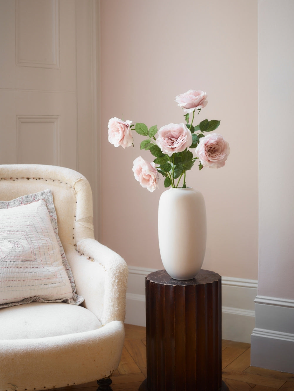 Faux roses stems in chiffon pink in a smooth cream vase on a dark wooden plinth next to an armchair