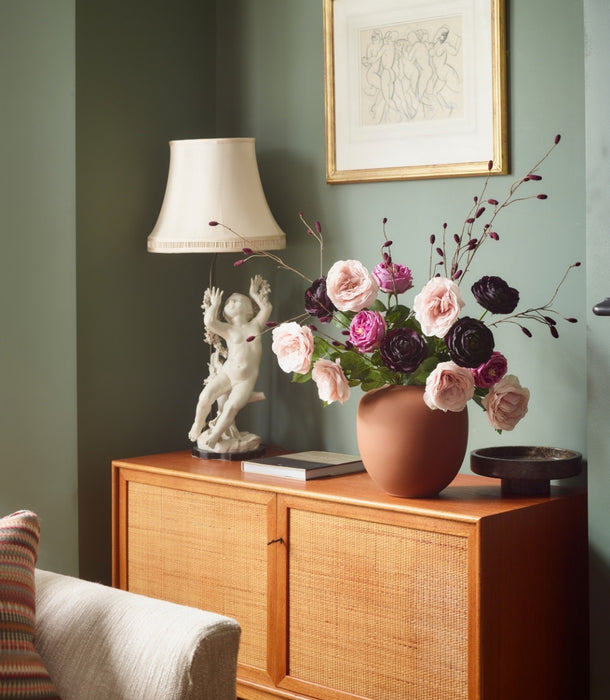bouquet of faux flowers including chiffon pink roses in a brown vase on a sideboard in a sage green living room  