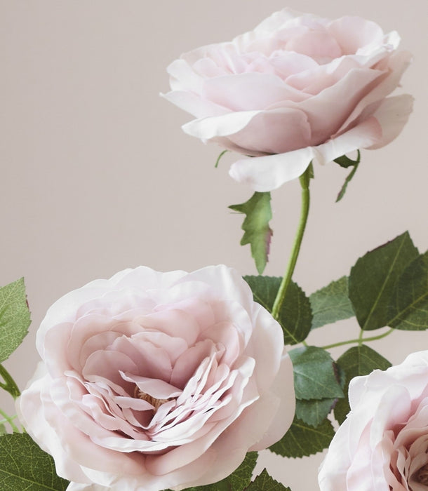 Three pale blush pink artificial roses with green stems and leaves against a soft beige background.