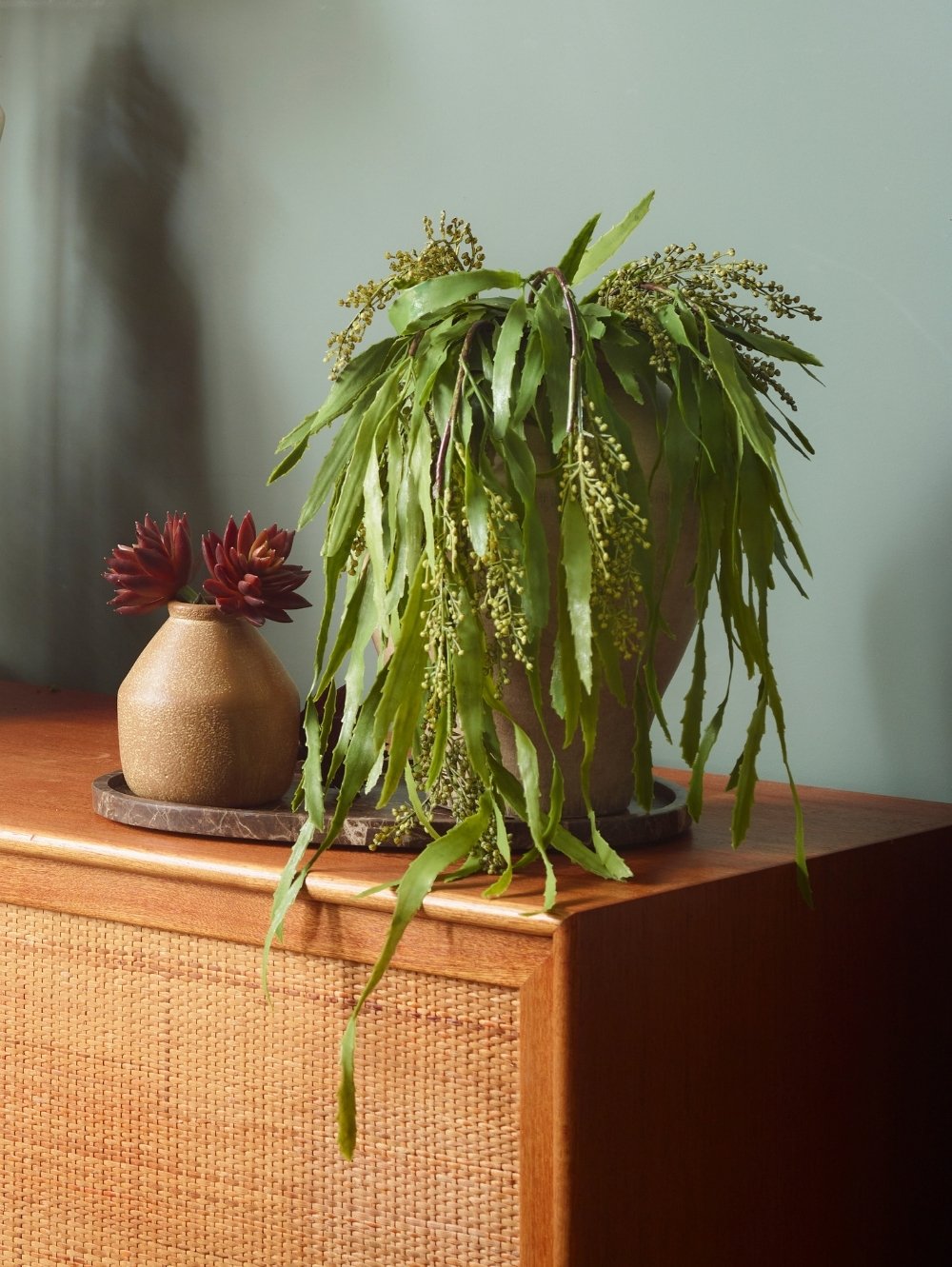 A green faux trailing berberis houseplant with narrow leaves and small clustered flowers sits in a dark pot on a wooden cabinet, next to a small beige vase with burgundy flowers against a muted green wall.
