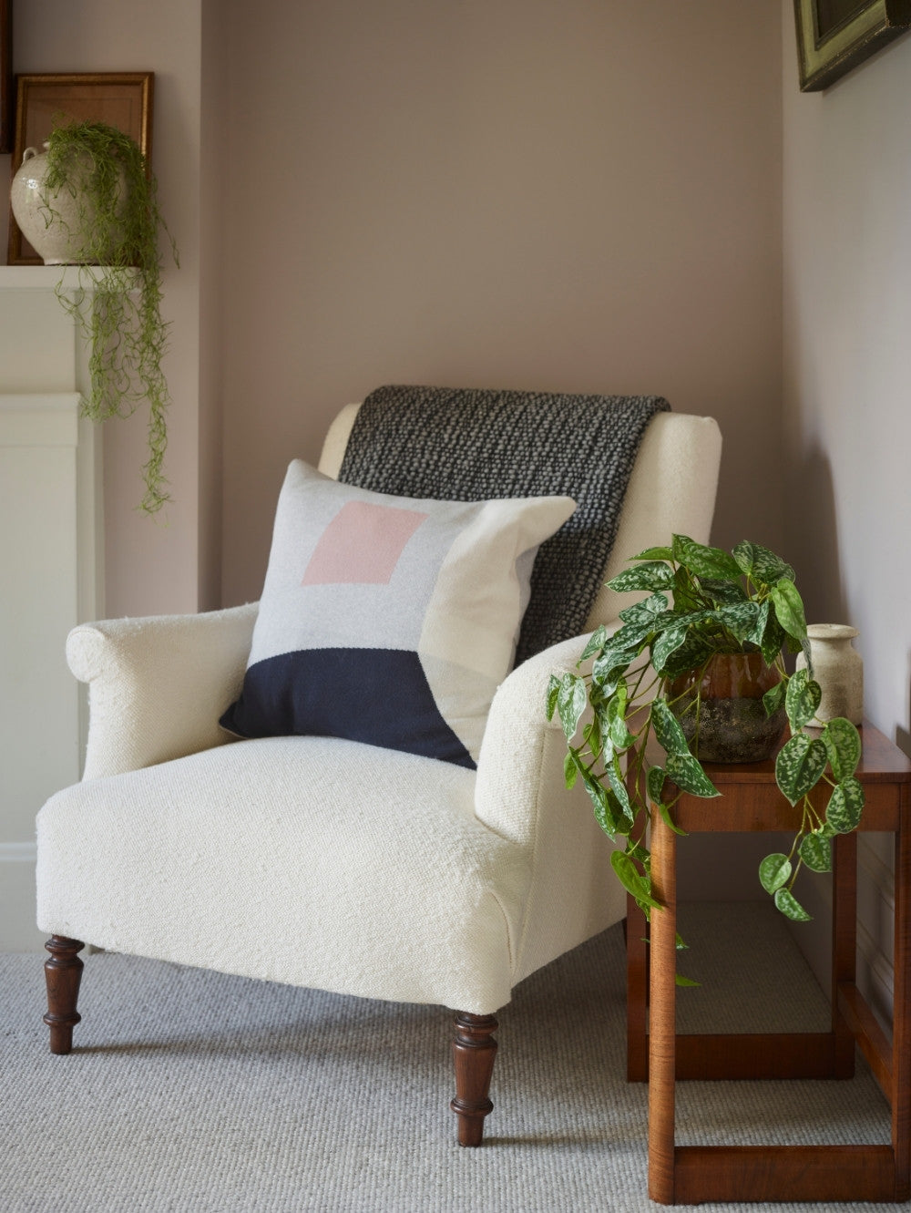 A cream bouclé armchair in a styled room corner, featuring a dark gray knit throw blanket draped over the back and a cream pillow with navy and light pink geometric blocks. A wooden side table with an artificial, green, trailing Scindapsus plant  sits next to the chair against a muted taupe wall.