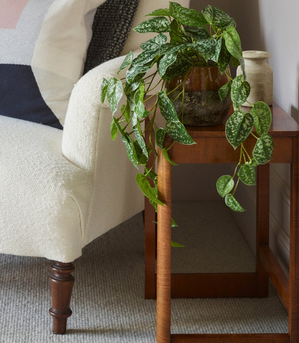 An artificial, green, trailing Scindapsus houseplant in a brown glass pot placed on a wooden side table next to a cream-colored armchair in a living room setting.