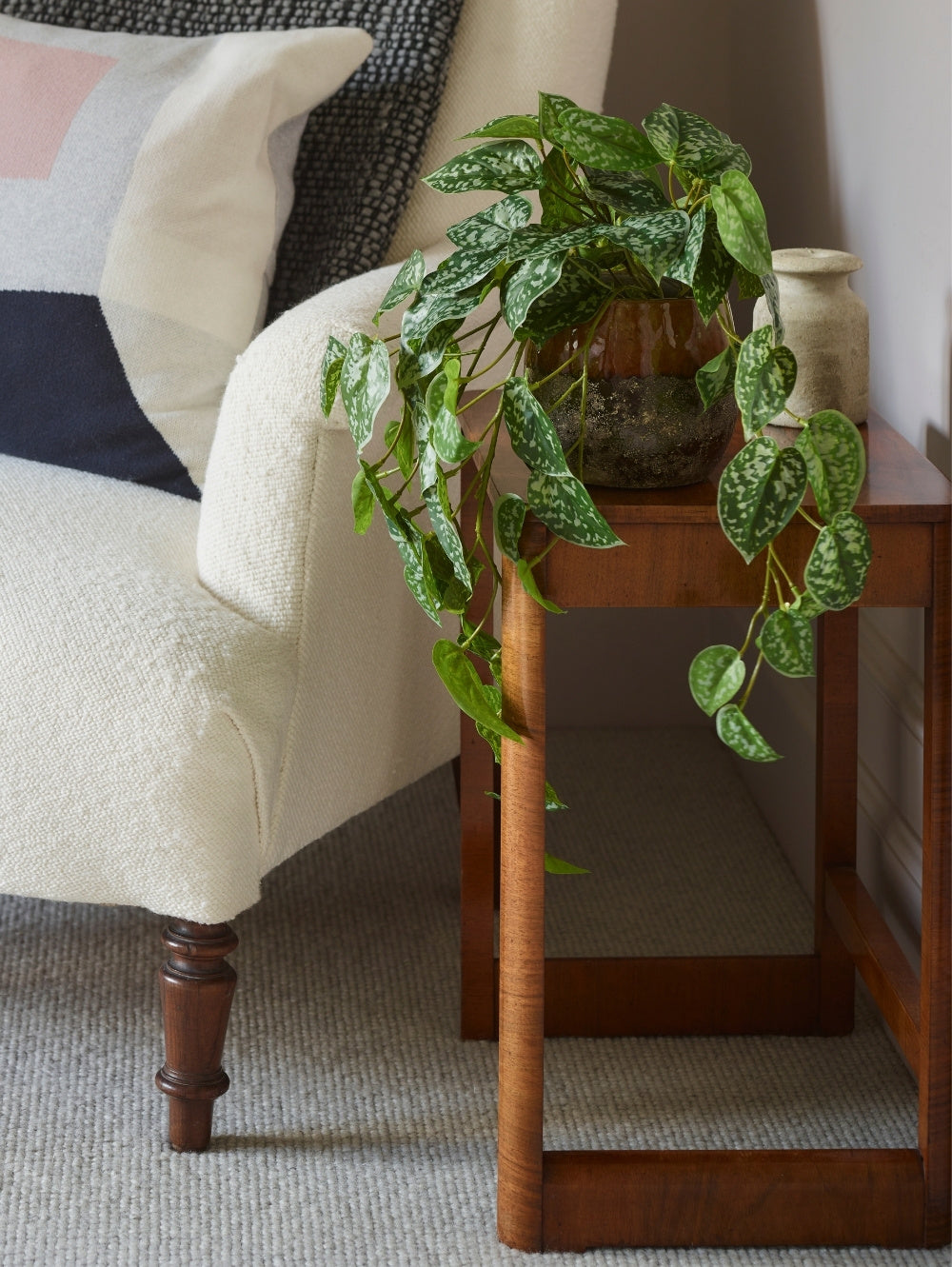 An artificial, green, trailing Scindapsus houseplant in a brown glass pot placed on a wooden side table next to a cream-colored armchair in a living room setting.
