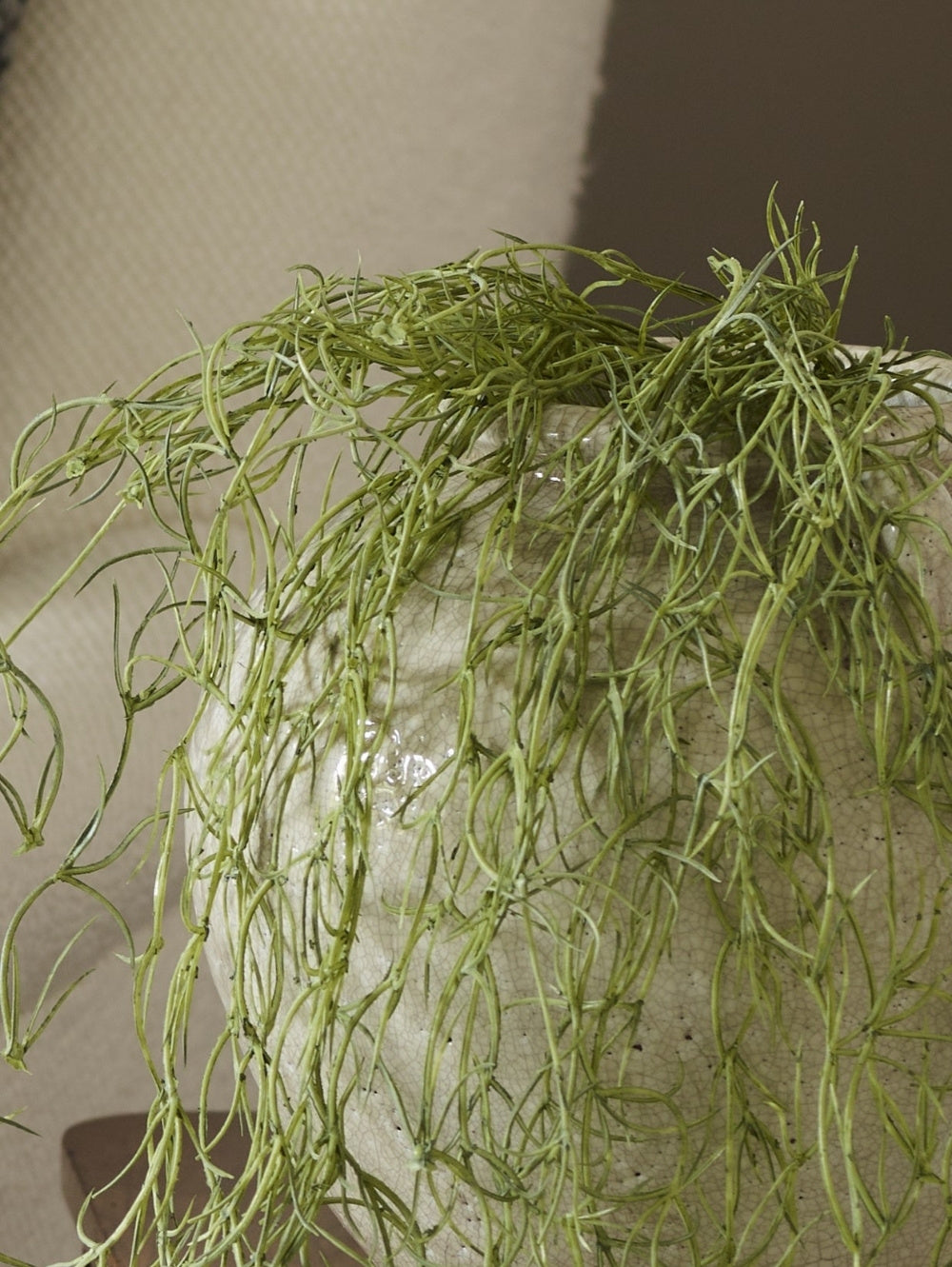 A close-up of an artificial trailing Spanish moss hanging plant with long, slender, thread-like leaves draped over a textured white ceramic pot against a neutral indoor background.
