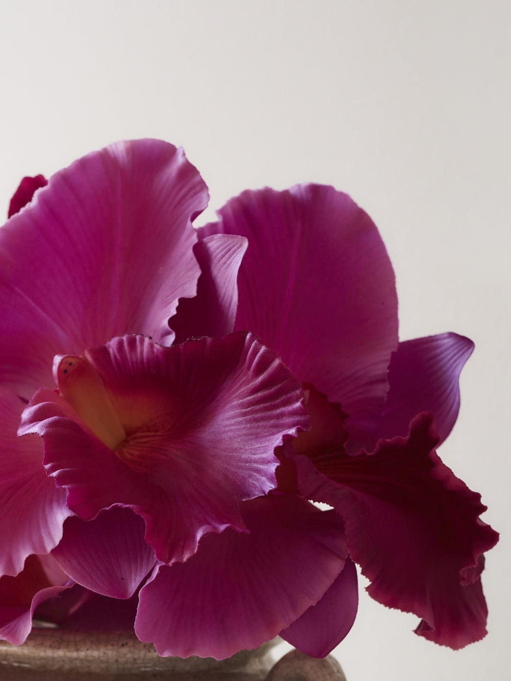 A close-up of an artificial blooming Vanda Orchid flower with large ruffled magenta-purple petals and a warm yellow-orange centre, photographed against a soft light grey background.