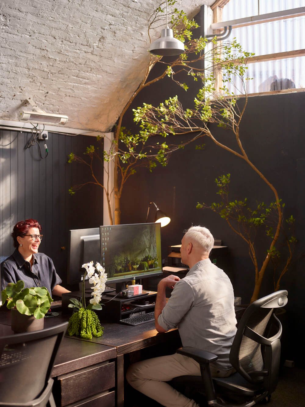 Two people working at a desk with computer monitors, surrounded by plants in an office setting.