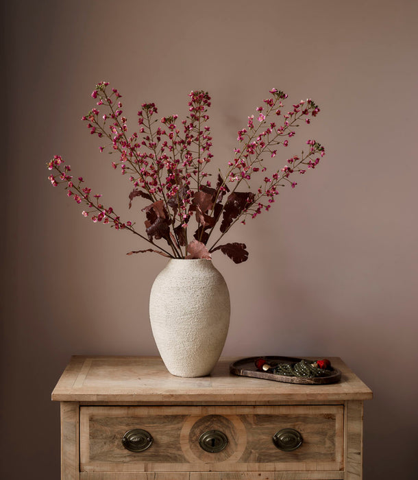 The Faux Primula Fandango, Pink - 85 cm stands in a textured white vase on a wooden dresser with ornate handles; a decorative tray with small items sits nearby, all set against a plain beige wall.