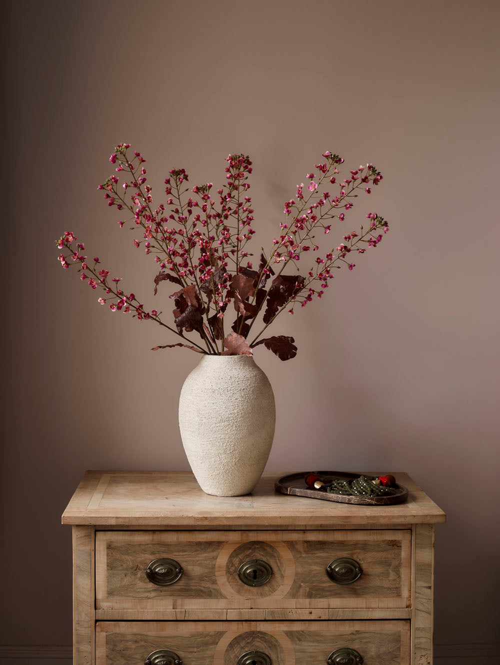 The Faux Primula Fandango, Pink - 85 cm stands in a textured white vase on a wooden dresser with ornate handles; a decorative tray with small items sits nearby, all set against a plain beige wall.