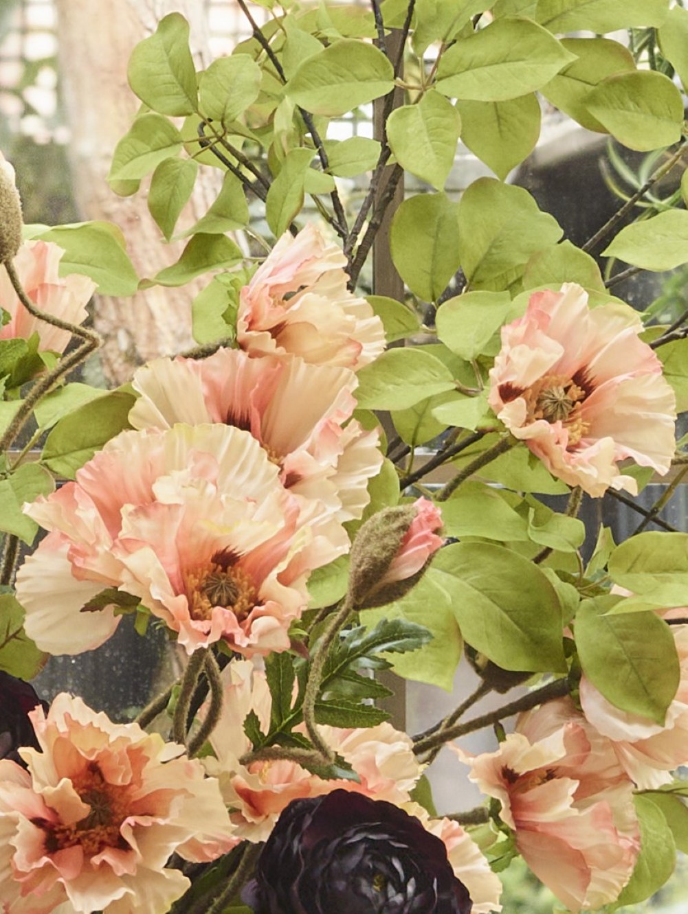 Close-up of faux coral poppies with delicate ruffled peach-coral petals and dark centres, paired with burgundy ranunculus and fresh green foliage.