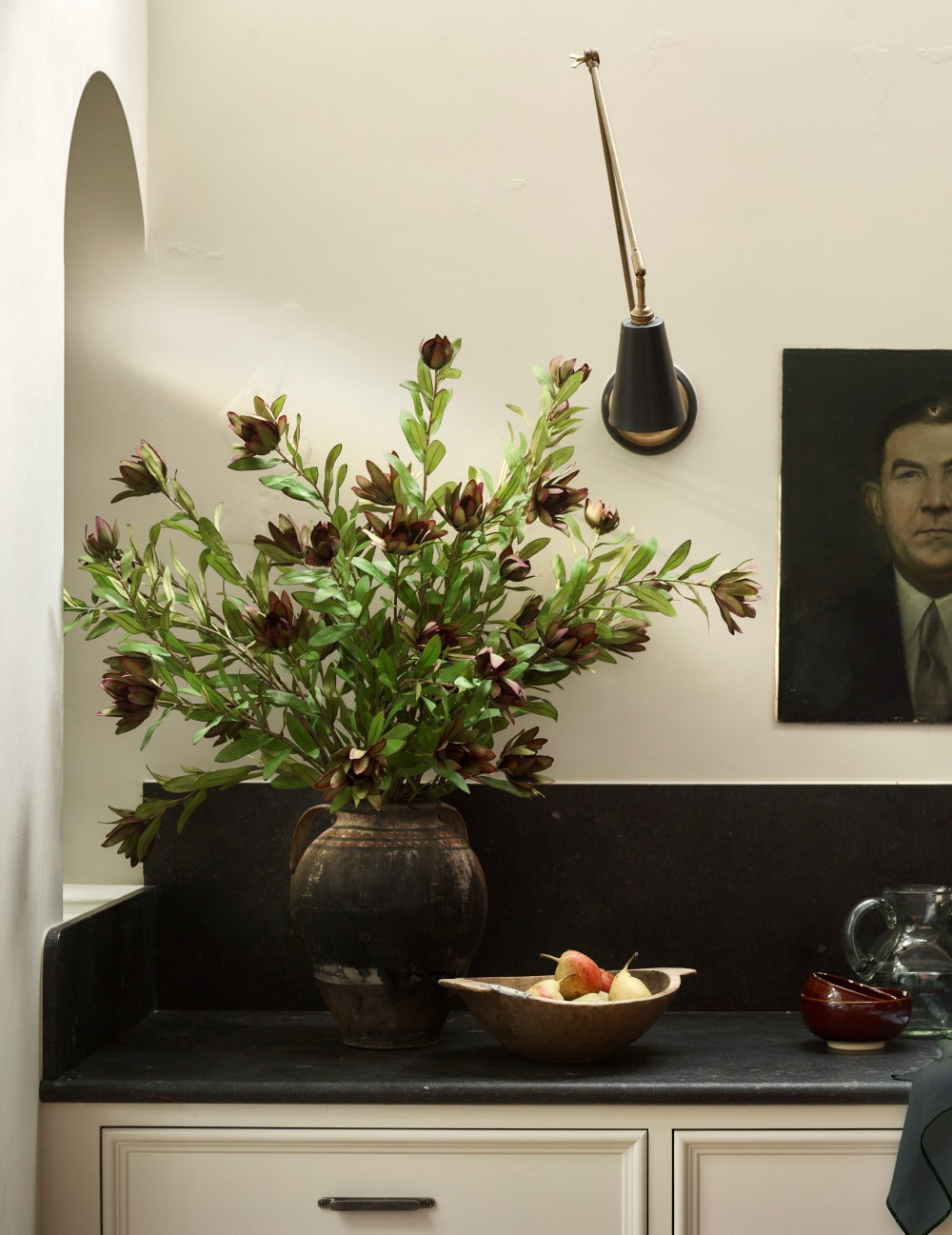 Green Faux Protea by Fake It Flowers, arranged in a dark ceramic vase on a black countertop, with burgundy flower heads and green foliage in a contemporary interior.