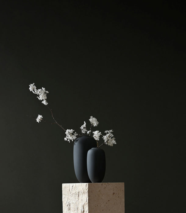 Two matte black 101 Copenhagen Sunao ceramic vases with delicate white floral sprigs, displayed on a textured beige stone pedestal against a solid dark charcoal background.