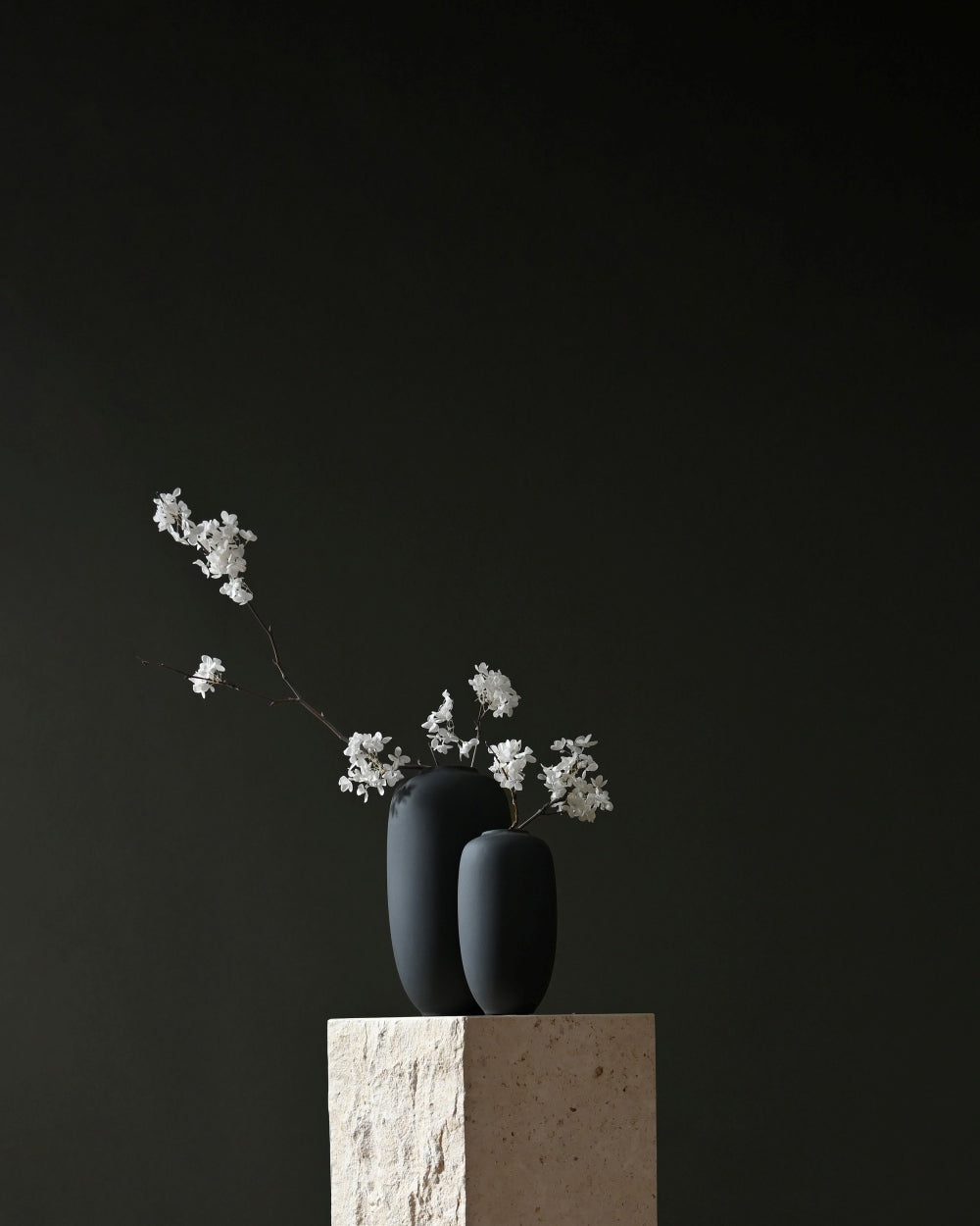 Two matte black 101 Copenhagen Sunao ceramic vases with delicate white floral sprigs, displayed on a textured beige stone pedestal against a solid dark charcoal background.