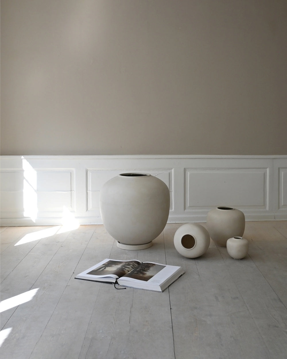 Sunao ceramic vases by 101 Copenhagen in Birch, arranged in varying sizes on a pale wooden floor beside an open book, against white panelled walls.