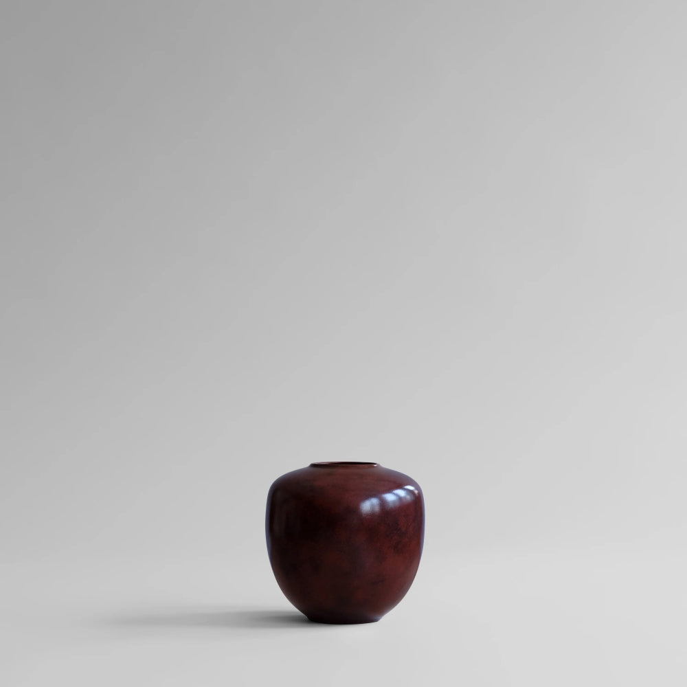 Sunao Vase Mini in glossy Maroon by 101 Copenhagen, styled with dark burgundy anthuriums, cream flowers and delicate branches, displayed on a wooden pedestal against white curtains.