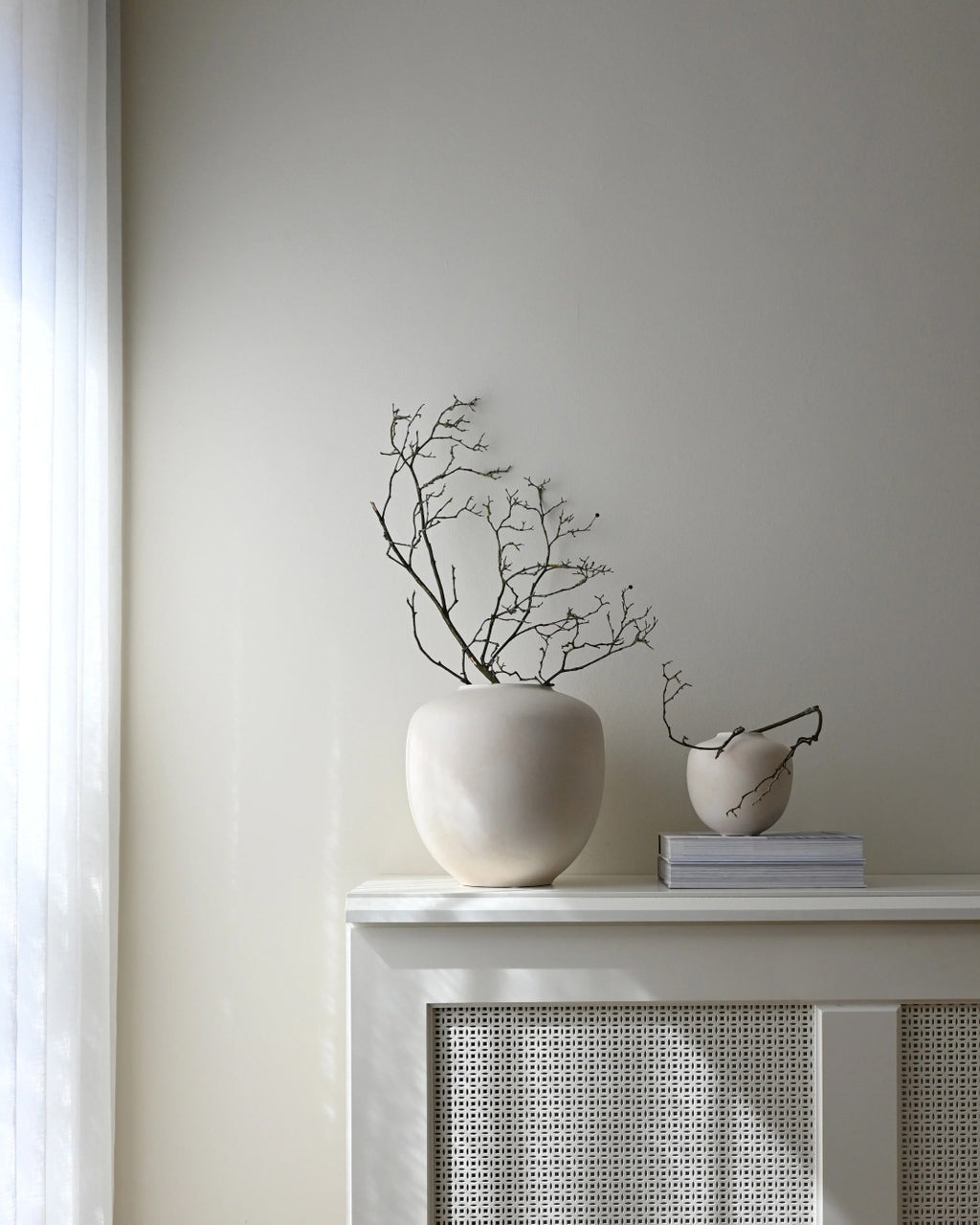 Sunao Vase Petit in Birch by 101 Copenhagen, a pale ceramic vase with soft rounded curves, styled with delicate bare branches on a white mantelpiece alongside a smaller ceramic vase and stacked books.