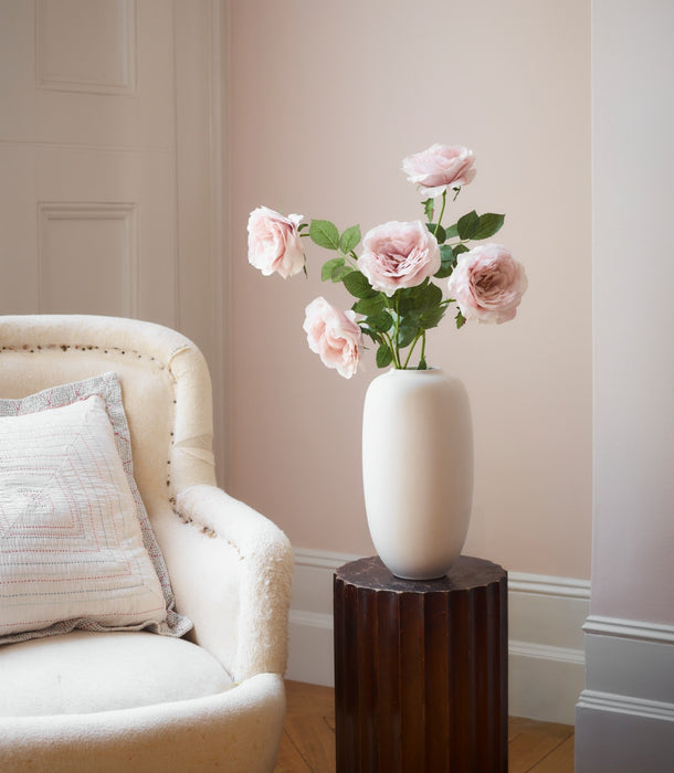 A birch 101 Copenhagen Sunao slim ceramic vase filled with chiffon pink roses with green leaves, displayed on a dark wood pedestal in a softly lit room. 