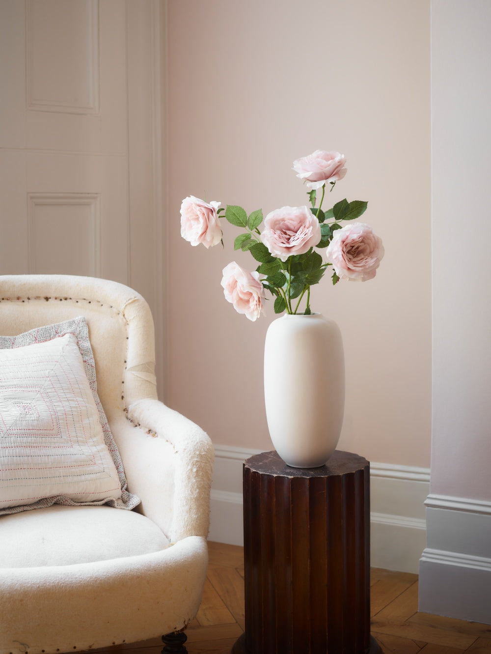 A birch 101 Copenhagen Sunao slim ceramic vase filled with chiffon pink roses with green leaves, displayed on a dark wood pedestal in a softly lit room. 