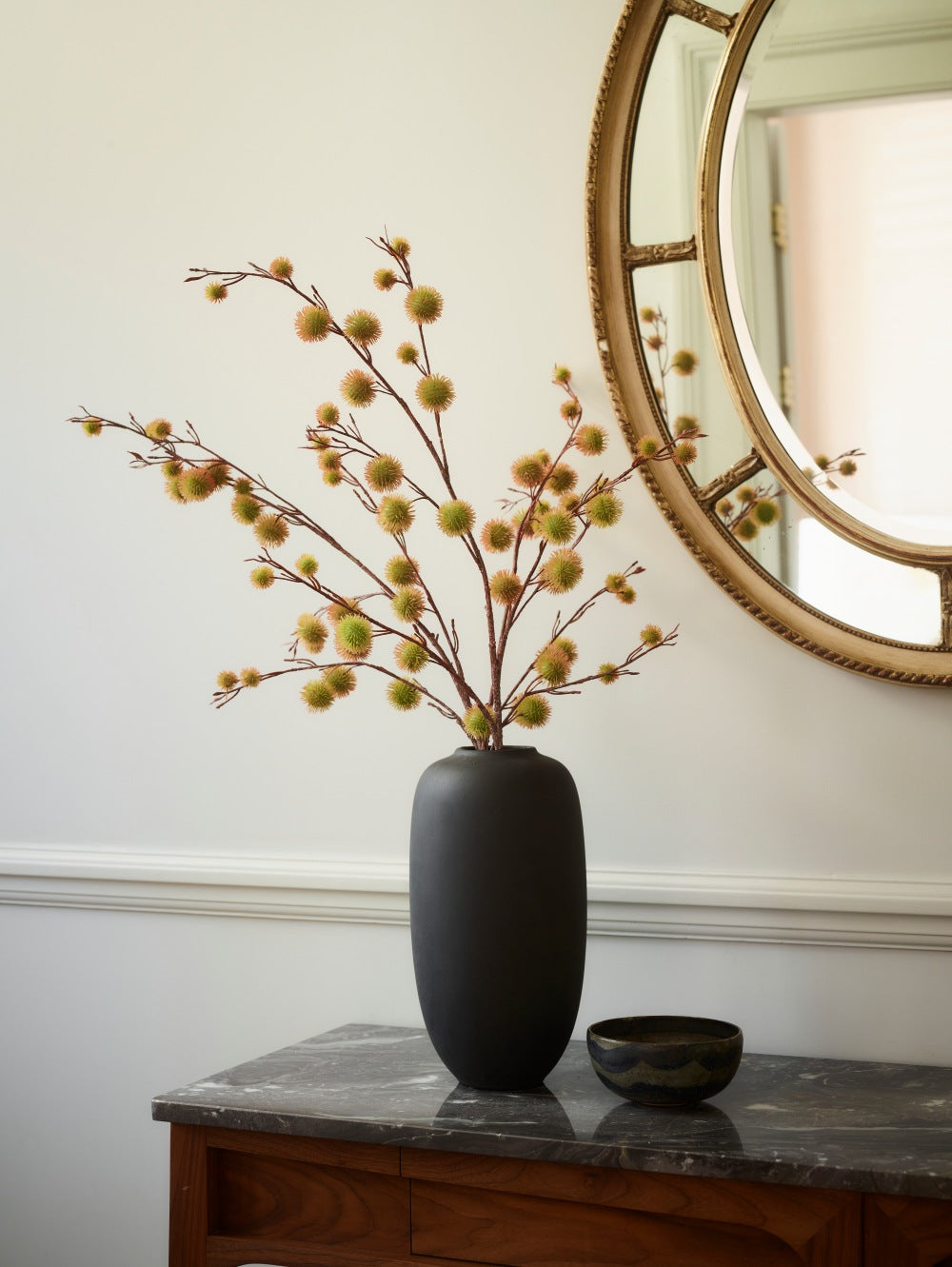 A matte black Sunao Vase in black ceramic vase filled with Chestnut yellow spray branches on a dark marble-topped console table beside a small matching bowl. 