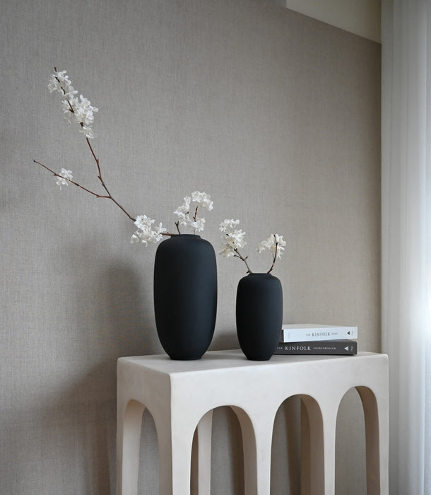 Two Sunao black vases in medium and small on a white console with white blossom sprays.