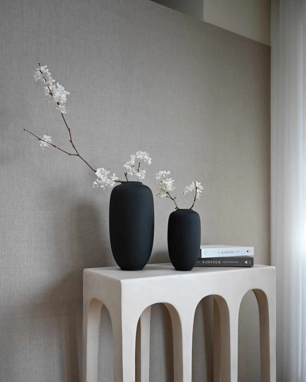 Two Sunao black vases in medium and small on a white console with white blossom sprays.