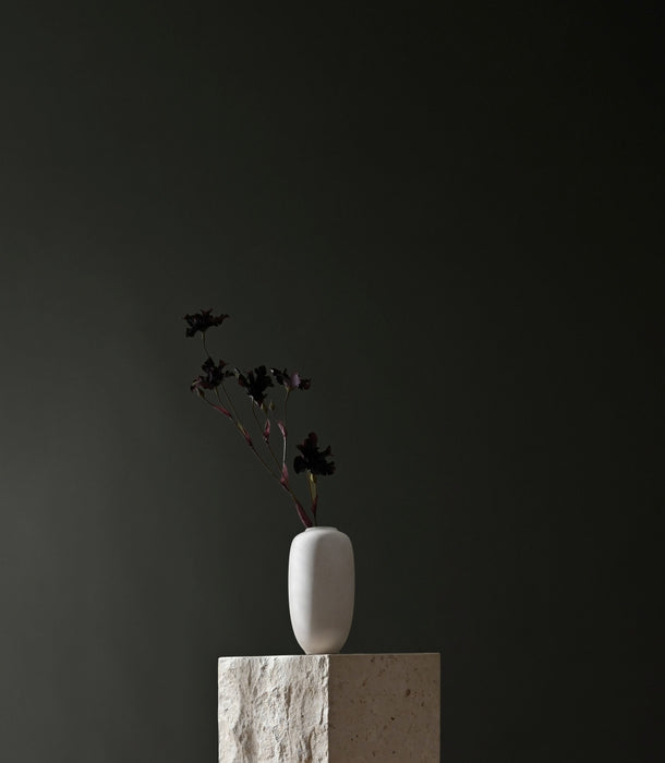 A minimalist white ceramic 101 Copenhagen Sunao vase with a small black floral arrangement, displayed on a textured stone pedestal against a dark charcoal background.