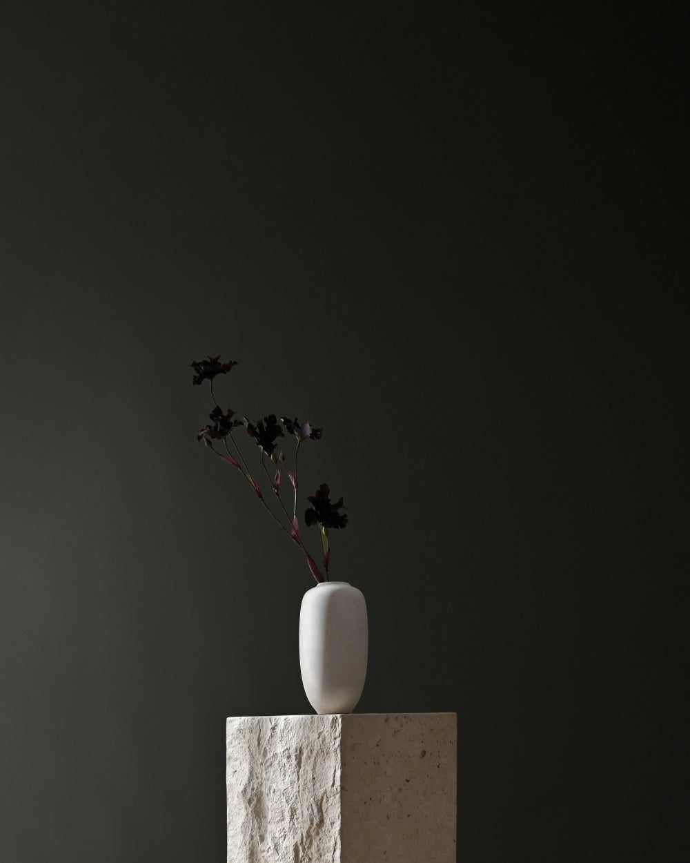 A minimalist white ceramic 101 Copenhagen Sunao vase with a small black floral arrangement, displayed on a textured stone pedestal against a dark charcoal background.
