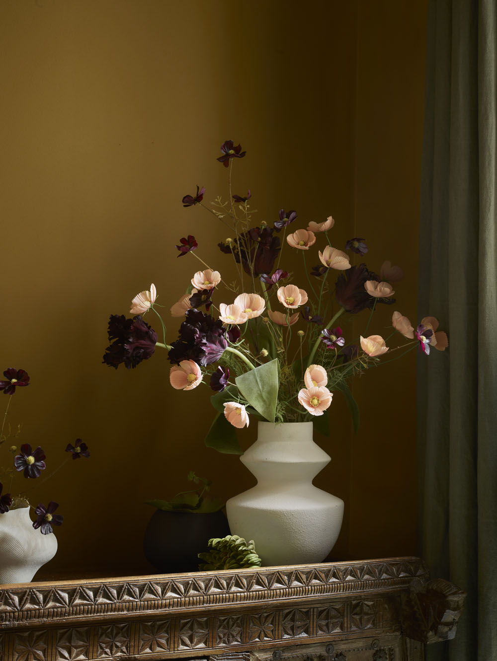 A white geometric vase displays Faux Parrot Tulip, Burgundy - 65cm, with ruffled petals in pink and deep purple on a carved wooden table against a mustard yellow wall, green curtain partly shown on the right.
