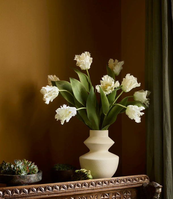 A 65cm White Faux Parrot Tulip with ruffled petals and green leaves sits on an ornate wooden table surrounded by potted succulents and moss, set against a mustard wall and dark green curtain.