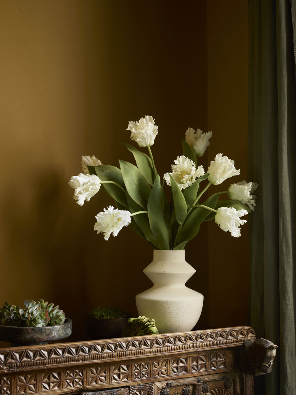 A 65cm White Faux Parrot Tulip with ruffled petals and green leaves sits on an ornate wooden table surrounded by potted succulents and moss, set against a mustard wall and dark green curtain.