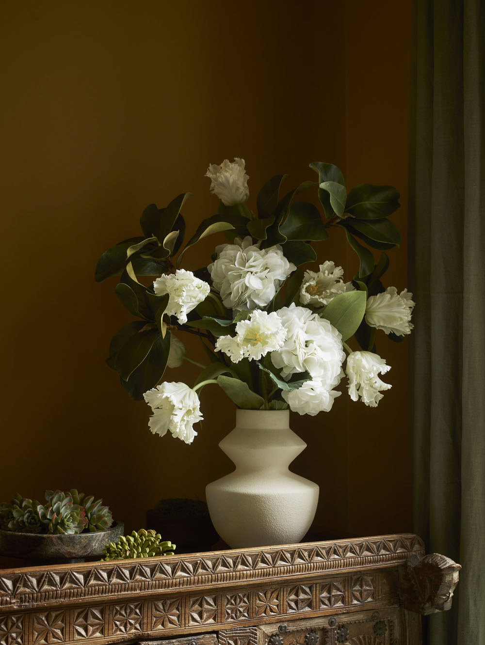 A cream-colored vase with Faux Parrot Tulip, White - 65cm displays ruffled white petals and lush green leaves on an ornate wooden table beside a succulent bowl, set against a warm brown wall and green curtain.