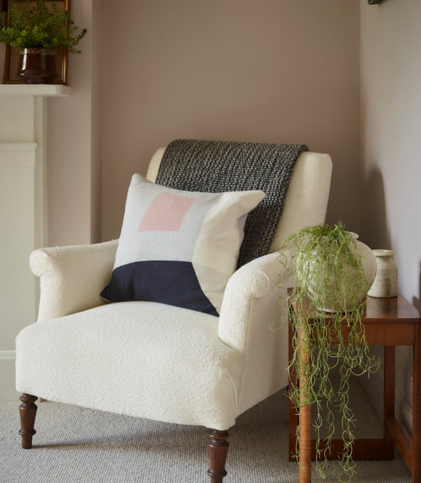 Our beautiful faux Spanish Moss stem flowing elegantly from a white urn planter next to a cream bouclé armchair in a styled room setting, featuring a dark gray knit throw blanket draped over the back