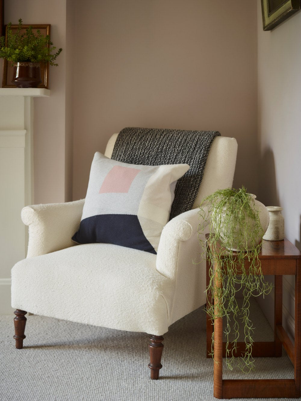 Our beautiful faux Spanish Moss stem flowing elegantly from a white urn planter next to a cream bouclé armchair in a styled room setting, featuring a dark gray knit throw blanket draped over the back