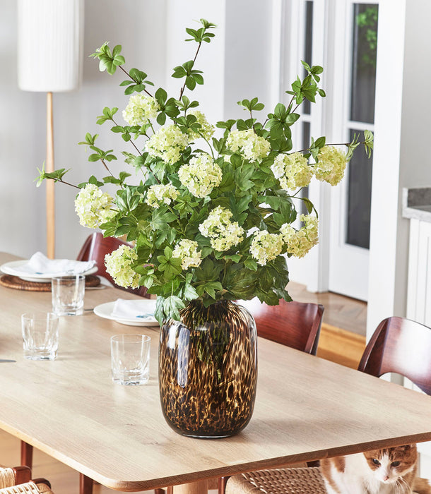 A Faux Viburnum, Pale Green - 78 cm arrangement in a brown patterned vase sits on a wooden dining table set with glasses and plates, while a cat lounges on a woven chair beneath the table.