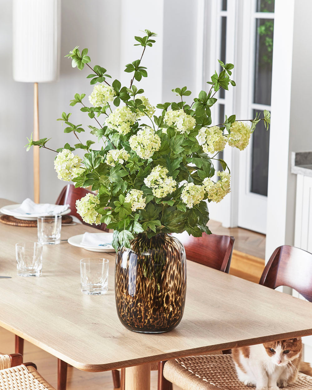 A Faux Viburnum, Pale Green - 78 cm arrangement in a brown patterned vase sits on a wooden dining table set with glasses and plates, while a cat lounges on a woven chair beneath the table.