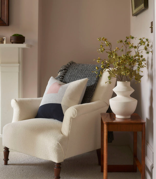 A cozy cream armchair with a patterned cushion and gray throw sits beside a wooden side table topped with a white vase holding Faux Winter Cherry, Green - 92 cm stems in a softly lit, neutral-toned living room.